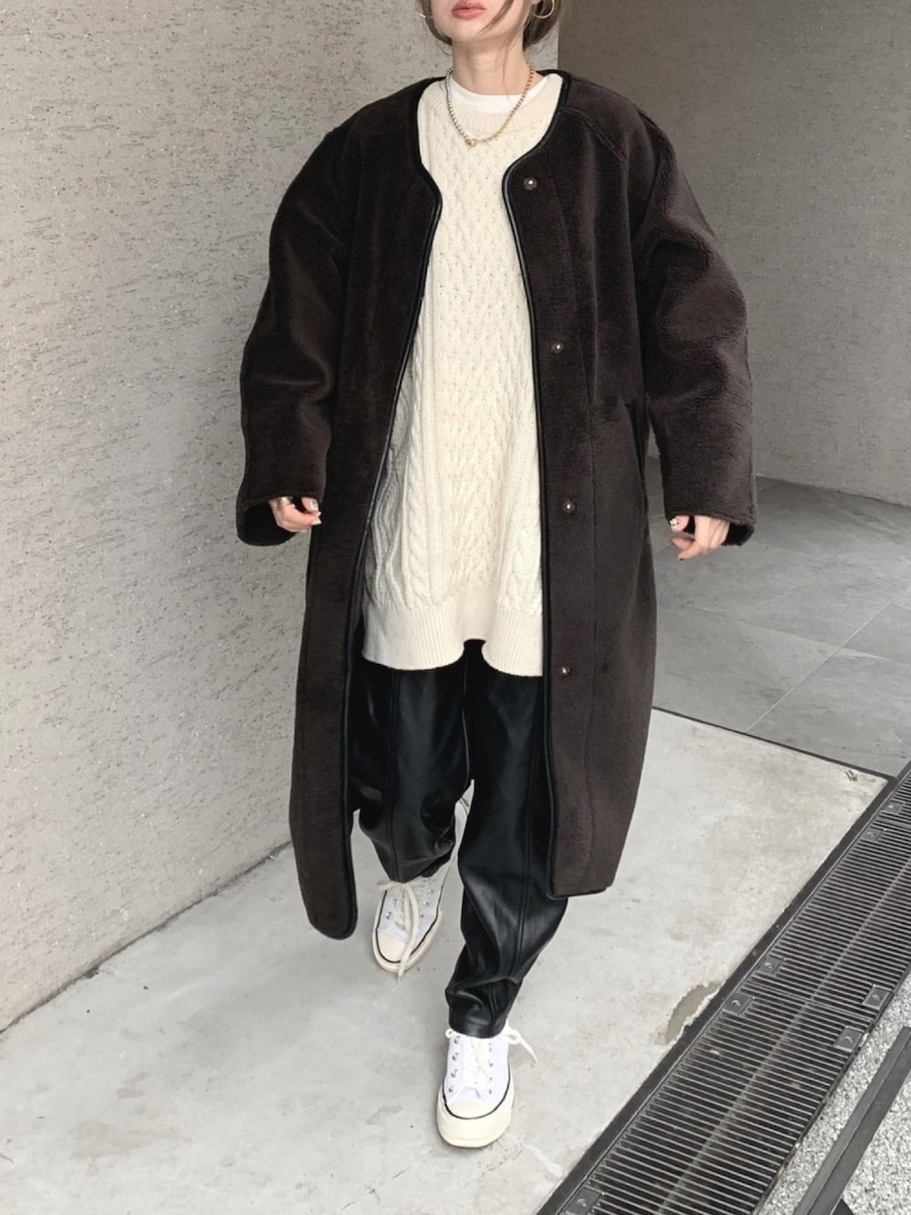 Woman in a long chocolate-brown teddy coat over a cream cable-knit sweater dress, black leather trousers and white sneakers