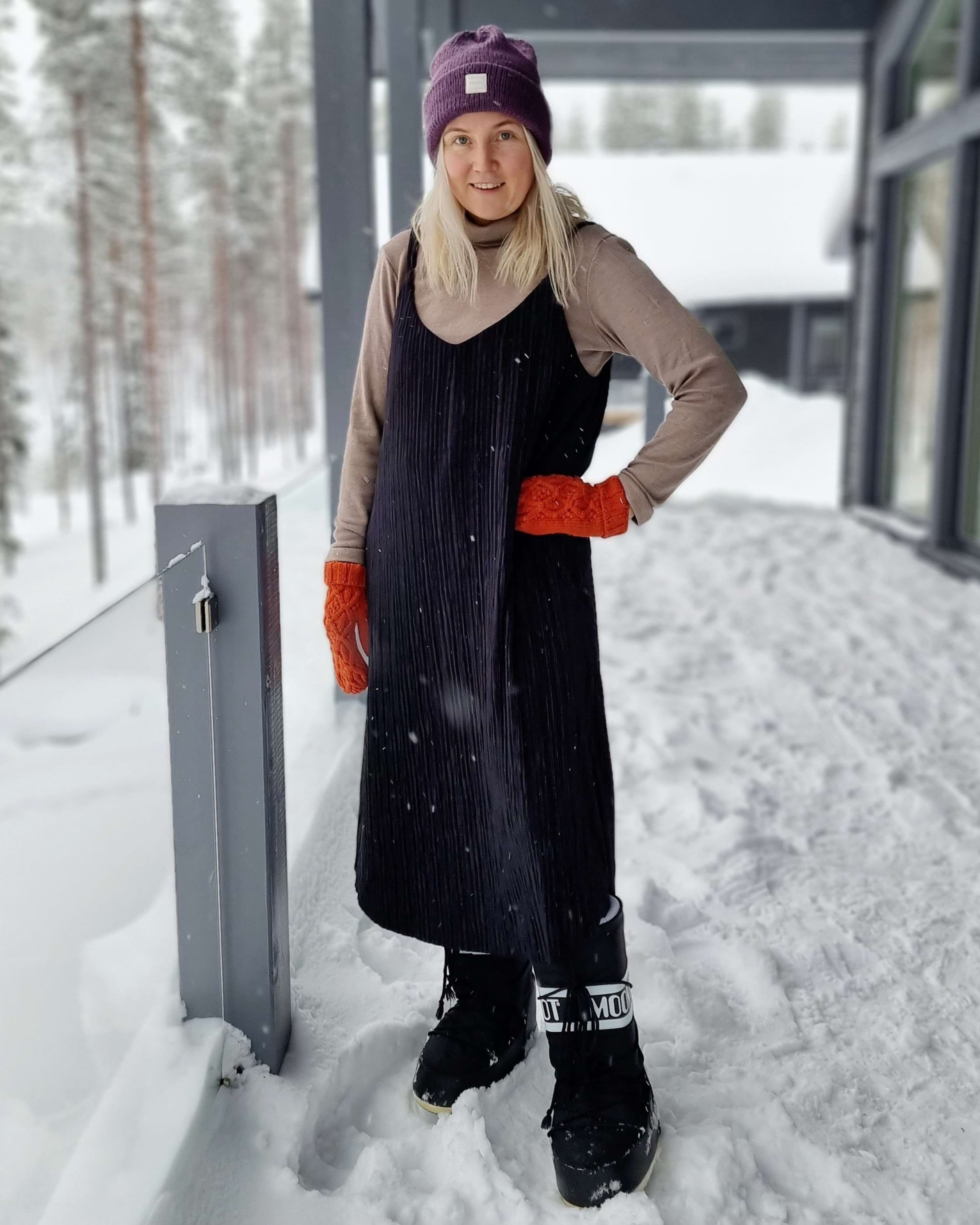 Woman in a black pleated dress, orange mittens and purple beanie wearing black moon boots in the snow