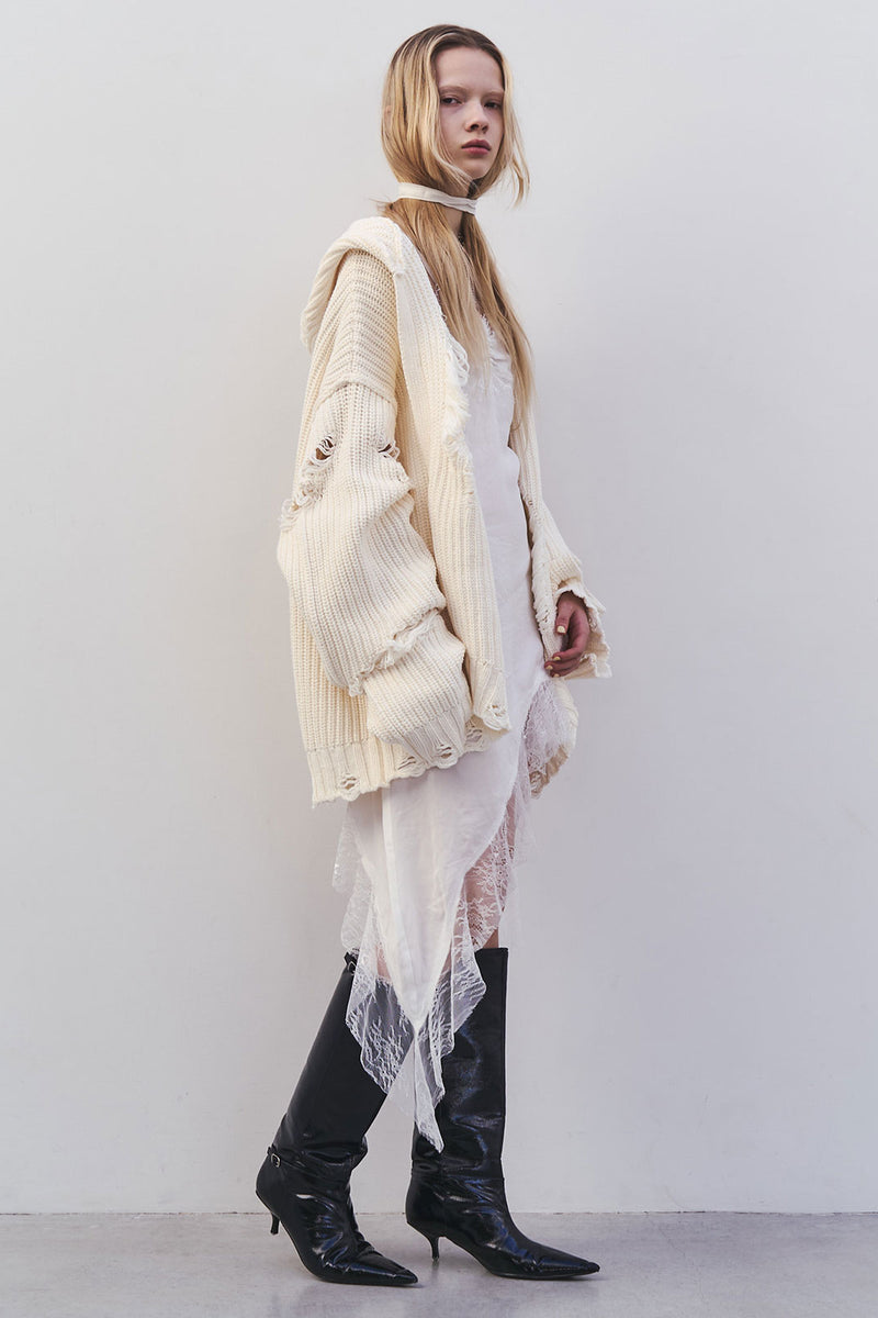 Woman wearing a distressed cream oversized cardigan over a white lace slip dress with black knee-high boots