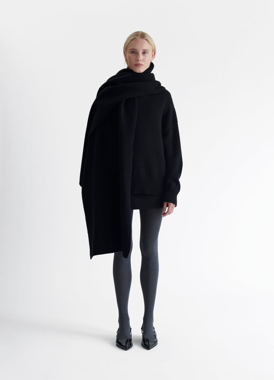 Woman in an all-black base of turtleneck sweater, tights and heels wrapped in an oversized black felted blanket scarf