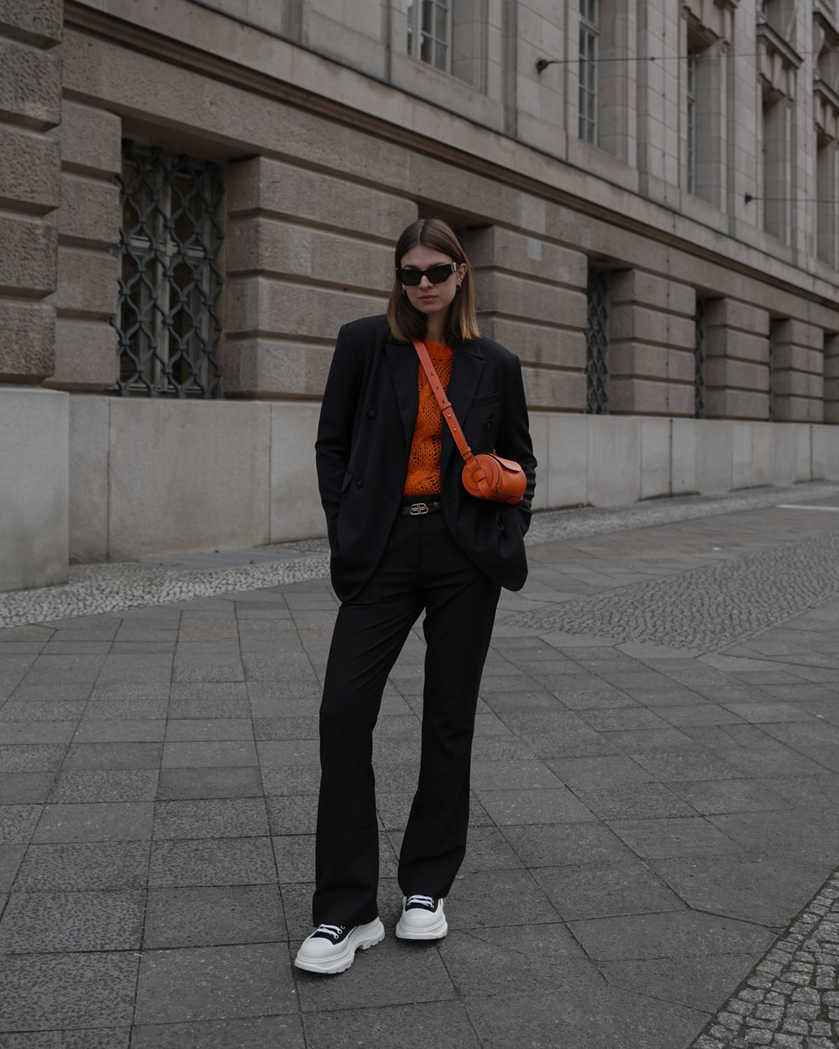 Woman in a chunky orange knit sweater, black blazer and black trousers with sneakers
