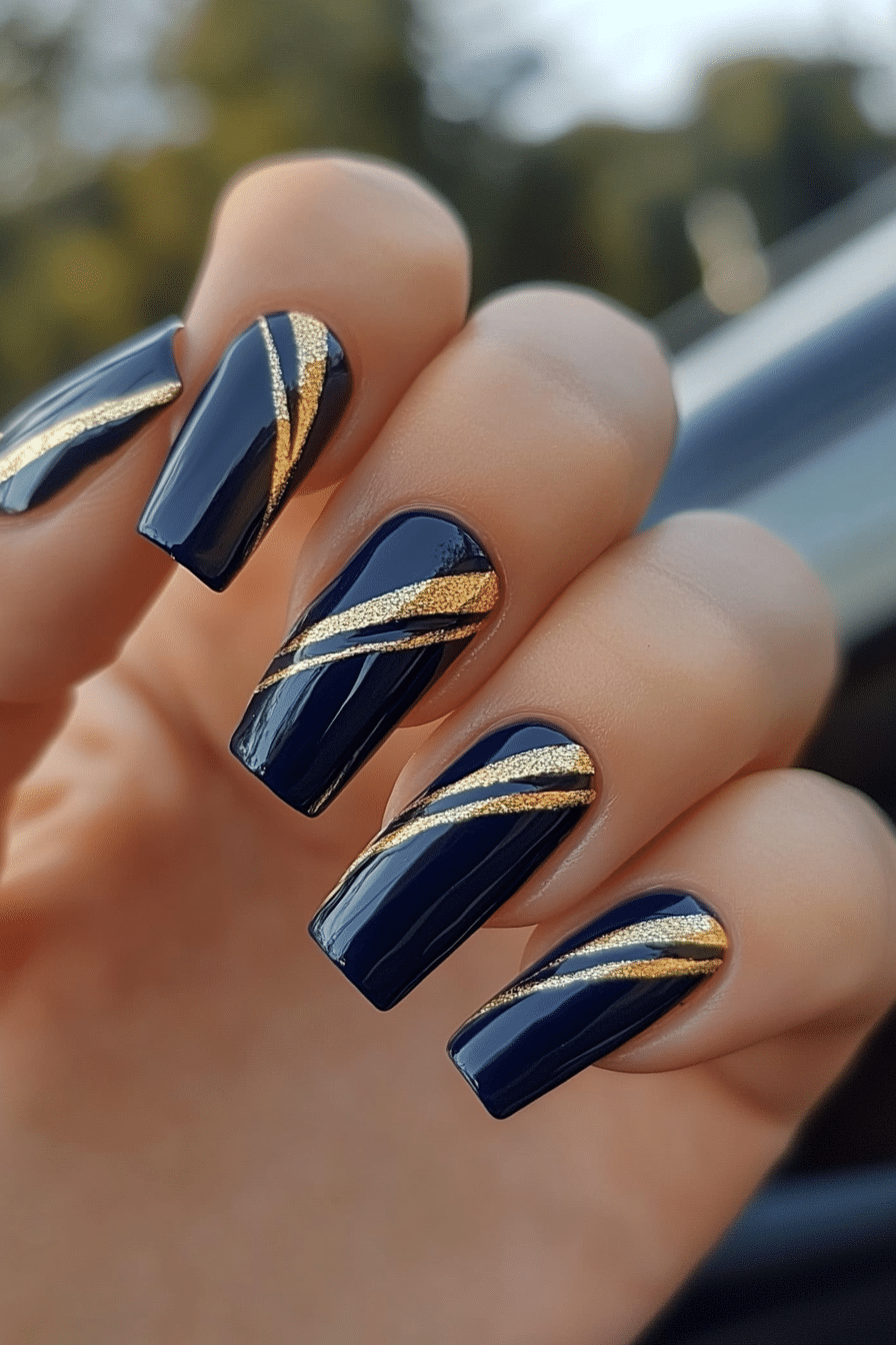 Long square navy nails with diagonal gold glitter gilded lines sweeping across each nail, photographed outdoors.