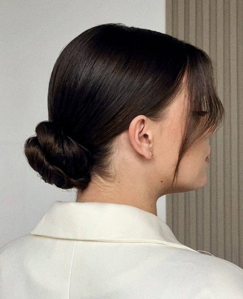 Sleek low knot bun with curtain bangs on dark brown hair, side profile. Note: credit page is a headband-hairstyles roundup; the photo itself