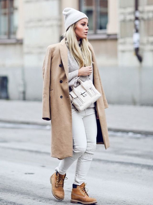 Woman in a camel coat, beige knit sweater, white skinny jeans, tan lace-up boots, gray beanie and a white structured bag walking on a city street