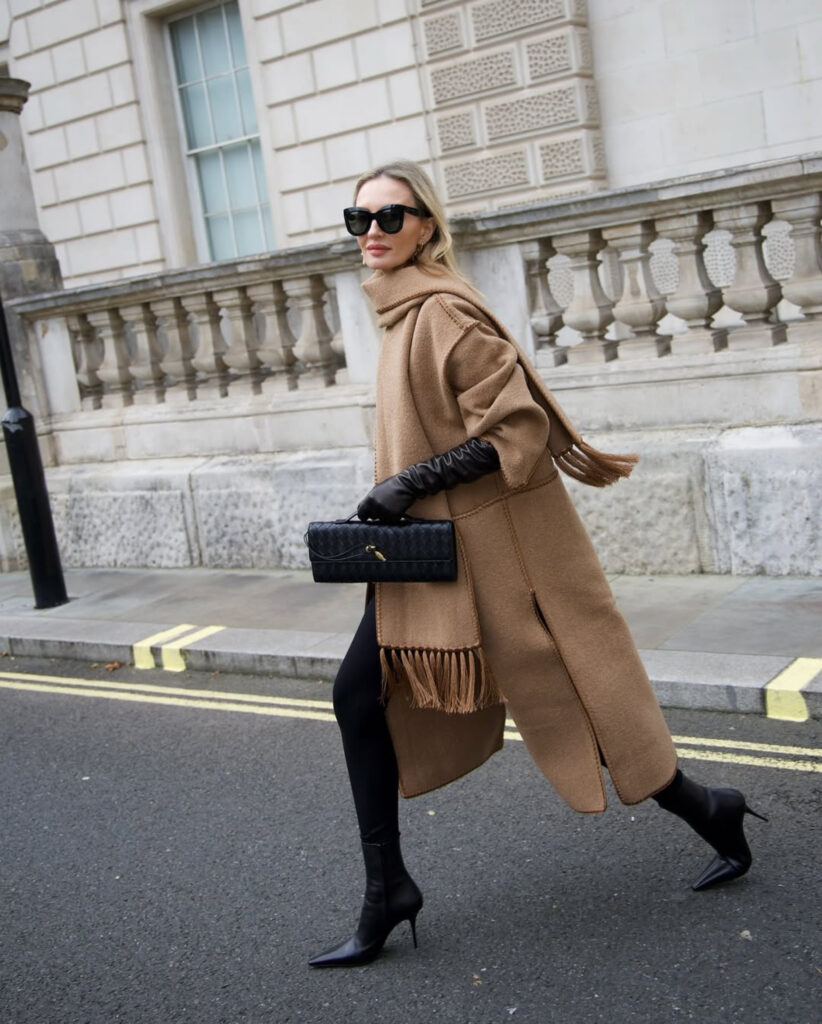 Woman in a camel coat with an attached fringed scarf, black leggings, long black gloves and stiletto ankle boots
