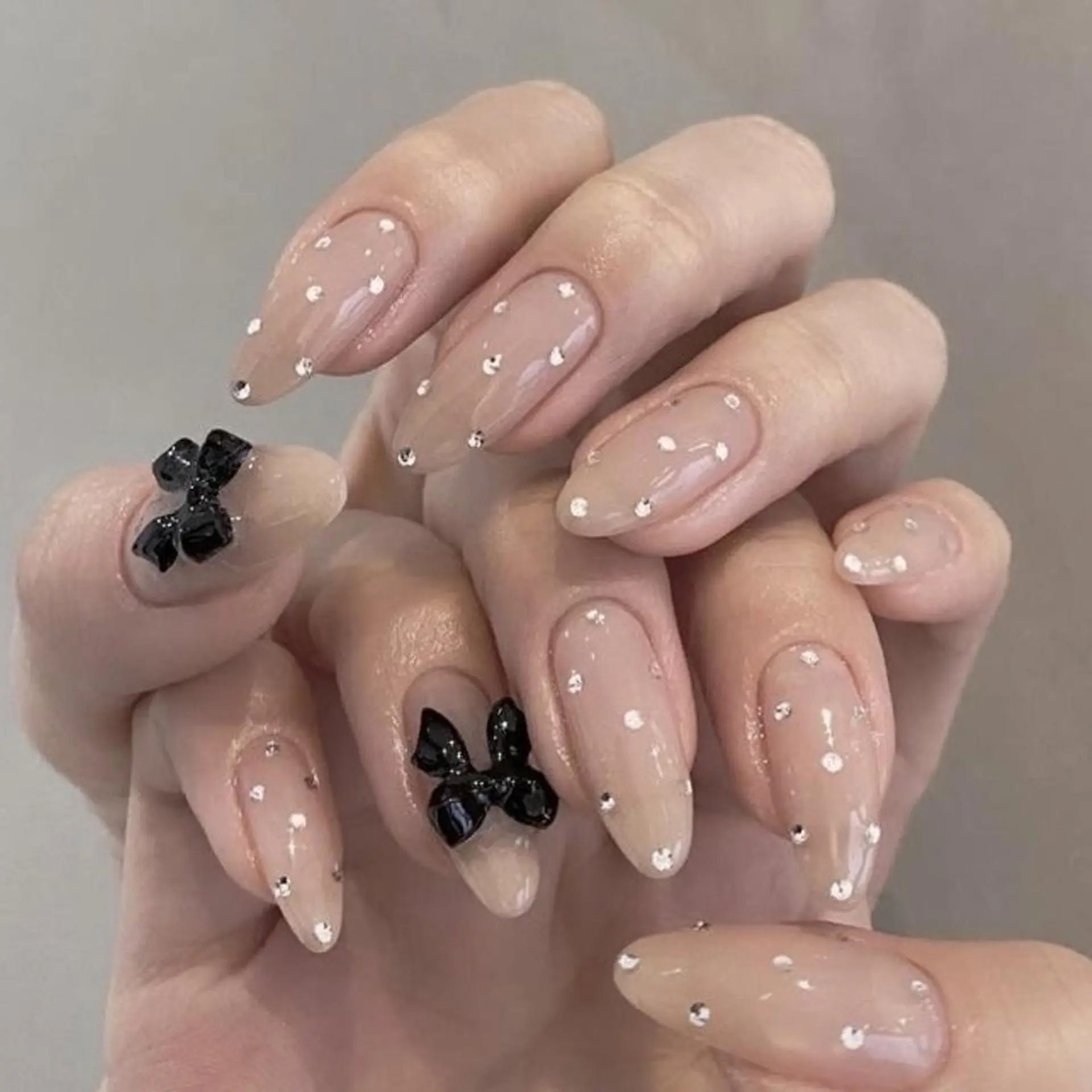 Medium almond nails in sheer nude with black 3D bow charms and scattered tiny rhinestones