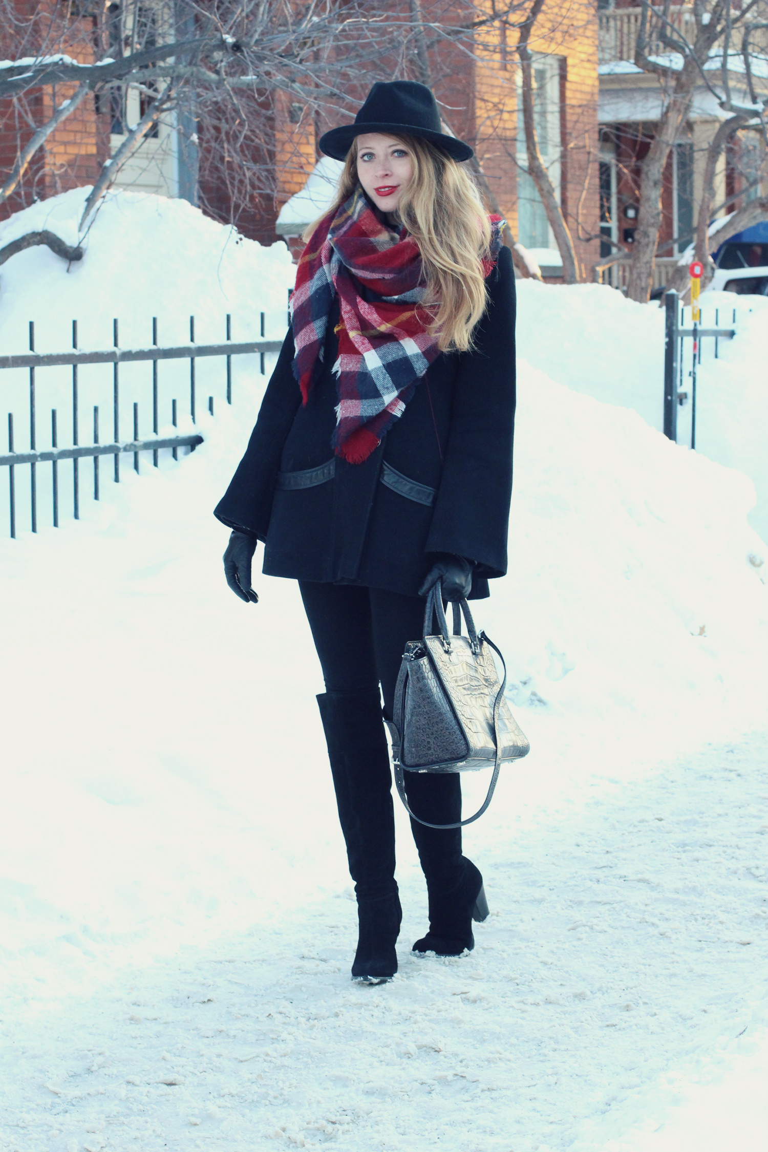 Woman in a black coat with a red plaid blanket scarf, black fedora and over-the-knee boots in the snow