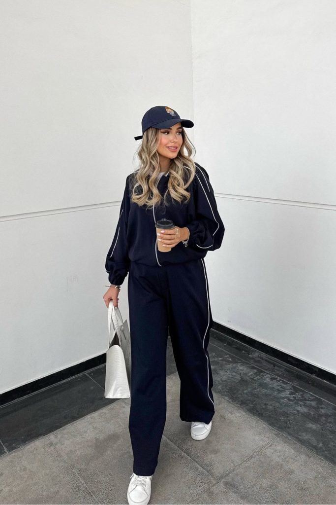 Woman in a navy piped tracksuit with a baseball cap, white sneakers and a tote bag
