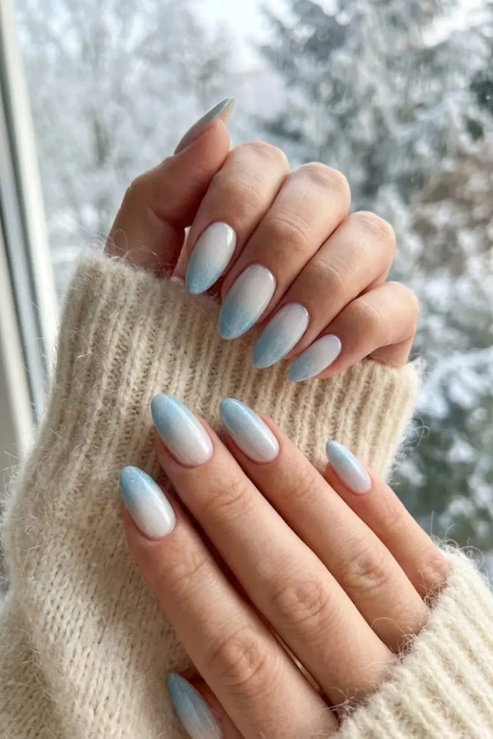 Almond nails with an ombre gradient from icy blue tips to white near the cuticle, hands in a cream knit sweater by a snowy window