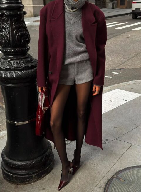 Woman in a burgundy wool coat and grey turtleneck sweater with grey shorts, tights and heels