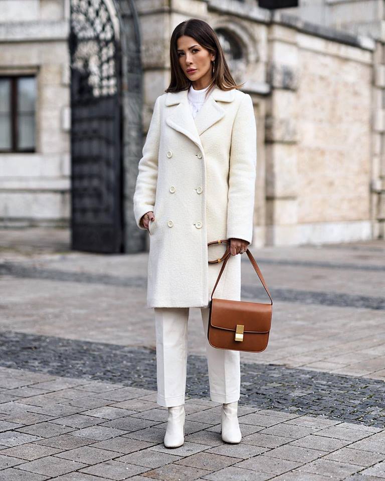 Cream double-breasted wool coat with white trousers, white boots, and a tan leather bag