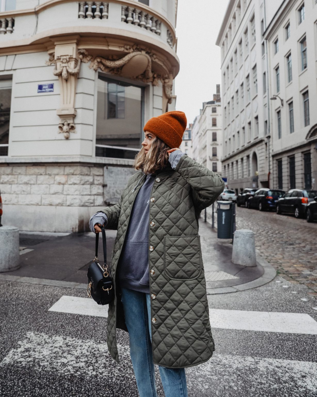 Woman on a Paris street wearing a long olive quilted coat over a grey hoodie, blue jeans, a rust knit beanie and carrying a black handbag