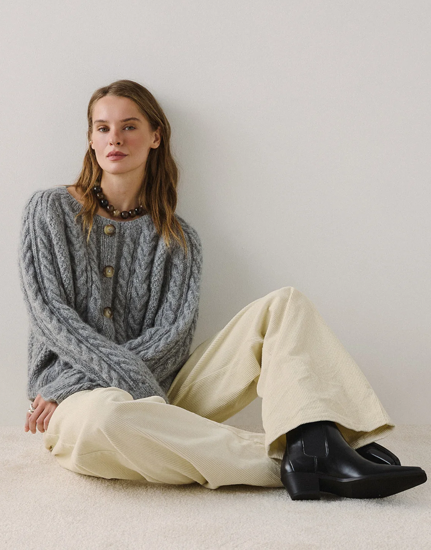 Woman seated wearing a grey cable-knit cardigan, cream corduroy wide-leg trousers and black Chelsea boots