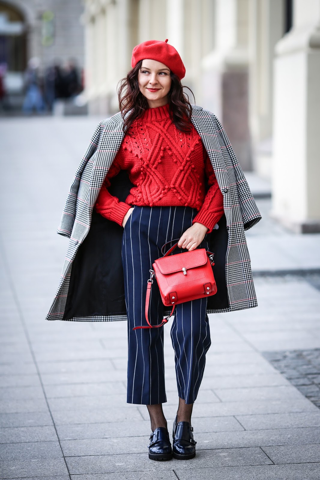 Woman in a red beret, red cable-knit sweater and navy pinstripe trousers with a houndstooth coat draped over shoulders
