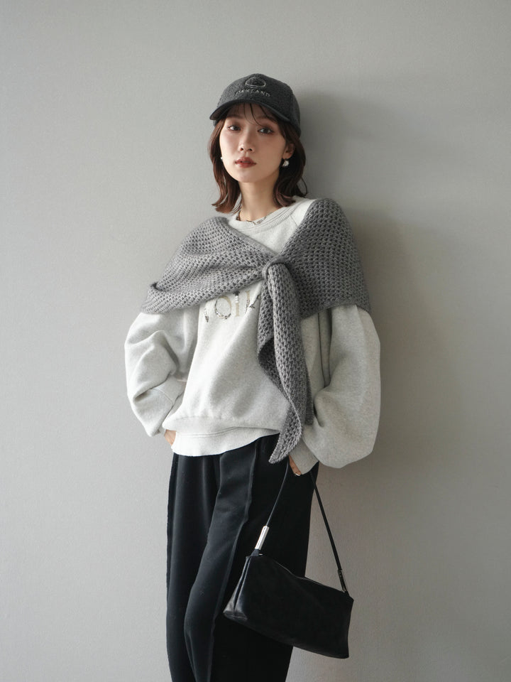 Woman wearing a gray knit shawl draped over a white sweatshirt with black wide trousers and a cap