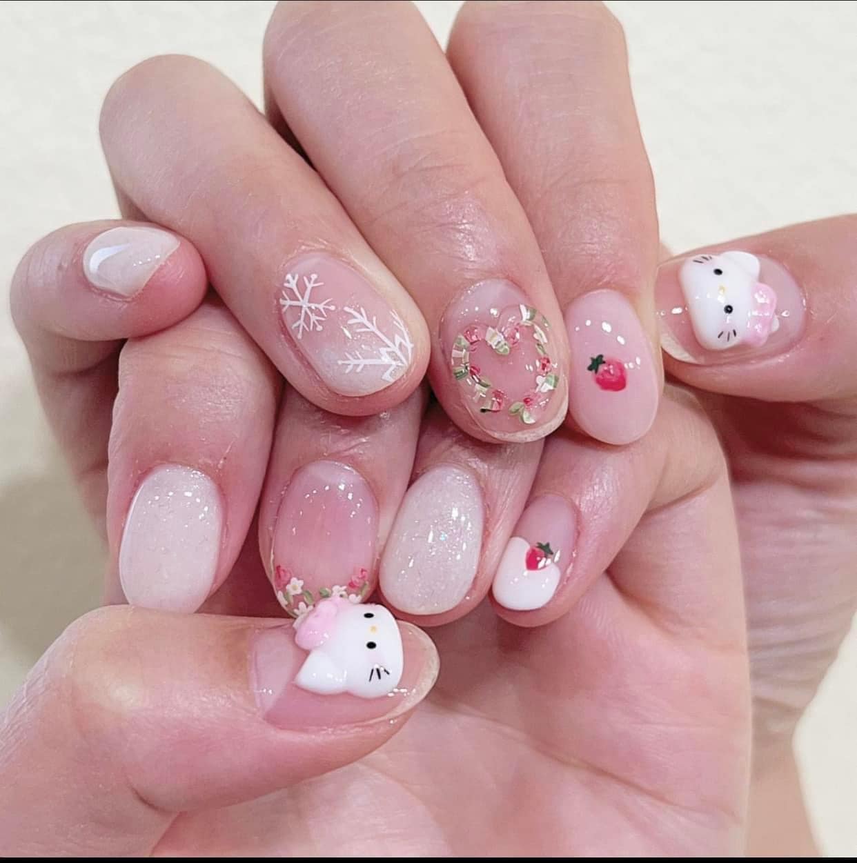 Short round natural nails in sheer pink with white snowflakes, a dried flower wreath, a small strawberry and Hello Kitty faces with pink bows