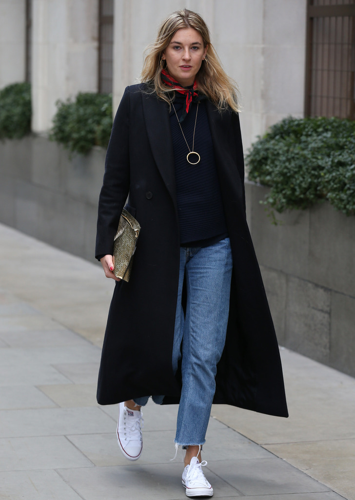 Woman in a black coat with a red neck scarf, navy knit sweater, blue jeans, and white sneakers
