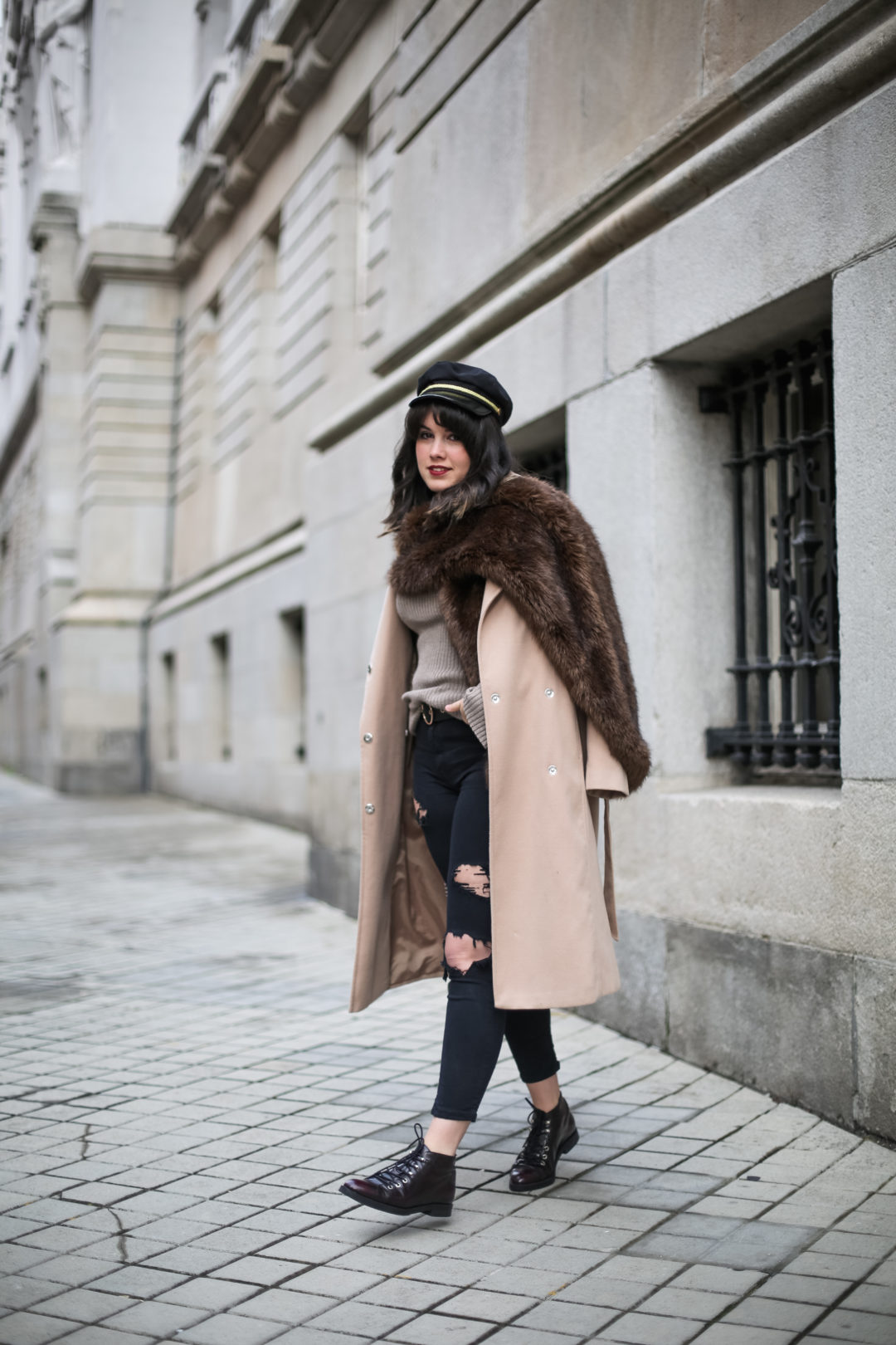 Woman in a brown faux fur stole over a camel wrap coat, beige knit, ripped jeans and lace-up boots