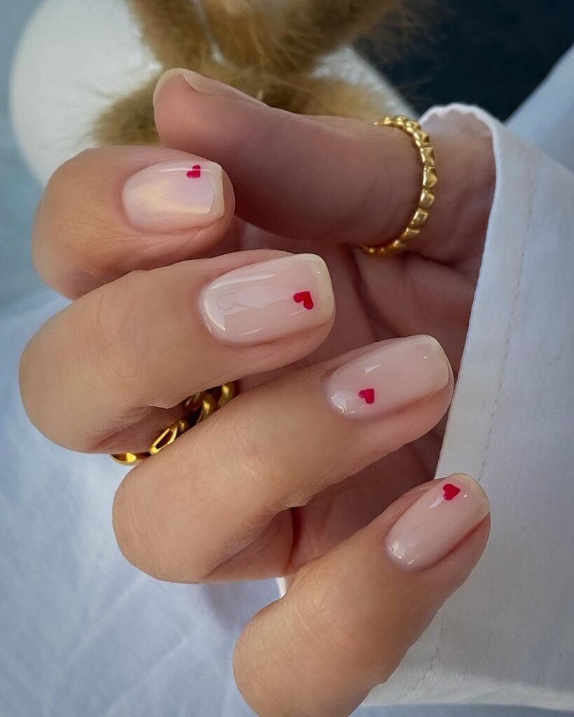 Short sheer nude nails, each with one tiny red heart near the tip.