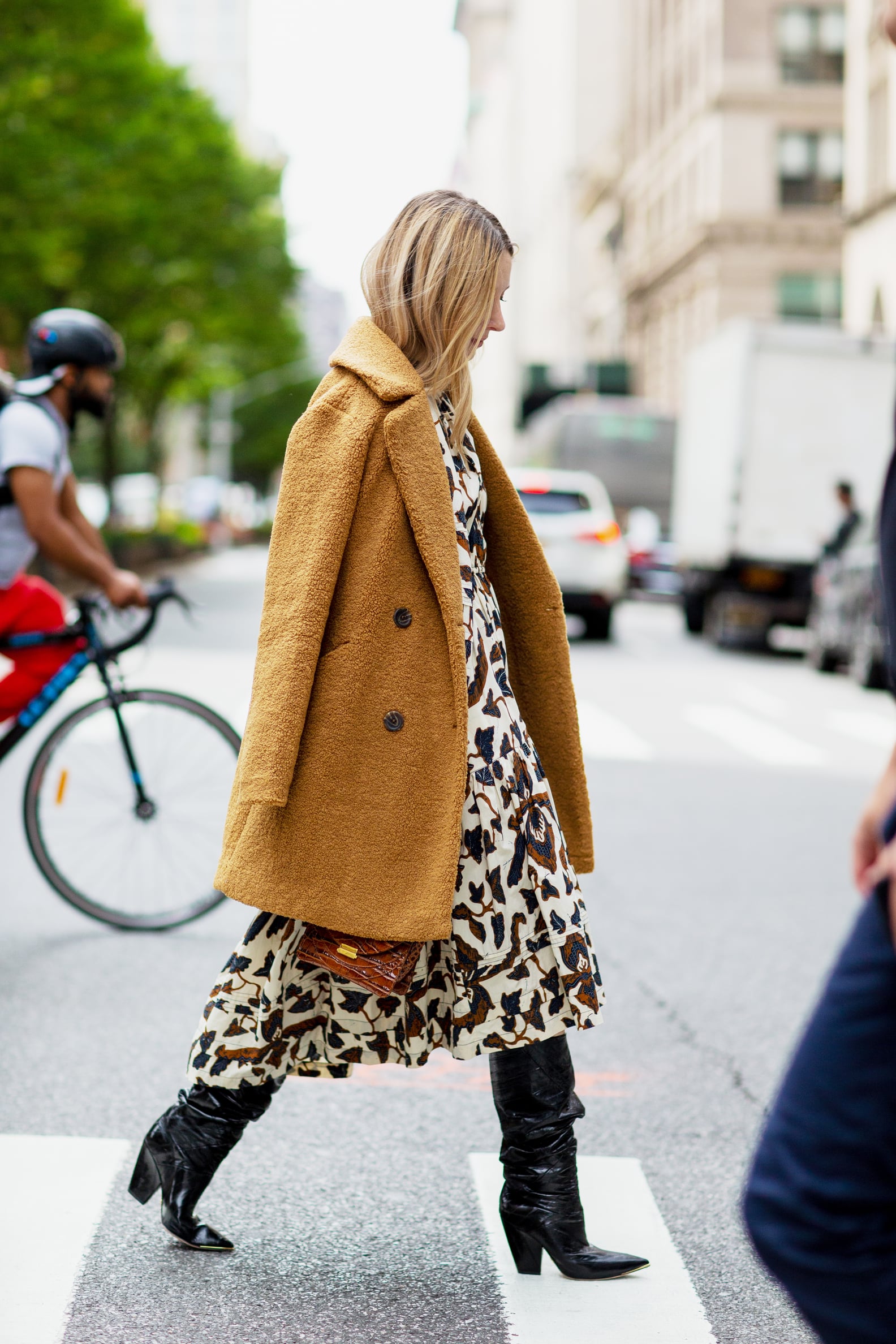 Woman in a camel teddy coat with a botanical-print midi dress and black slouch boots crossing the street