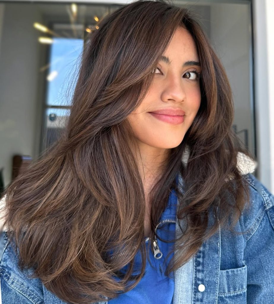 Woman with long dark brown wavy hair and a soft honey blonde money piece framing her face