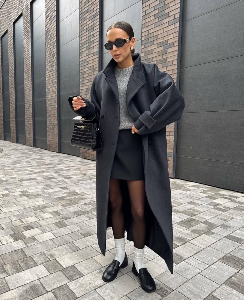 Woman in a long black coat with a grey sweater, black mini skirt, tights, white socks and loafers