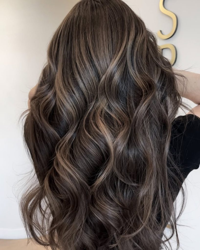 Woman with deep brunette wavy hair and cool mushroom brown face-framing highlights