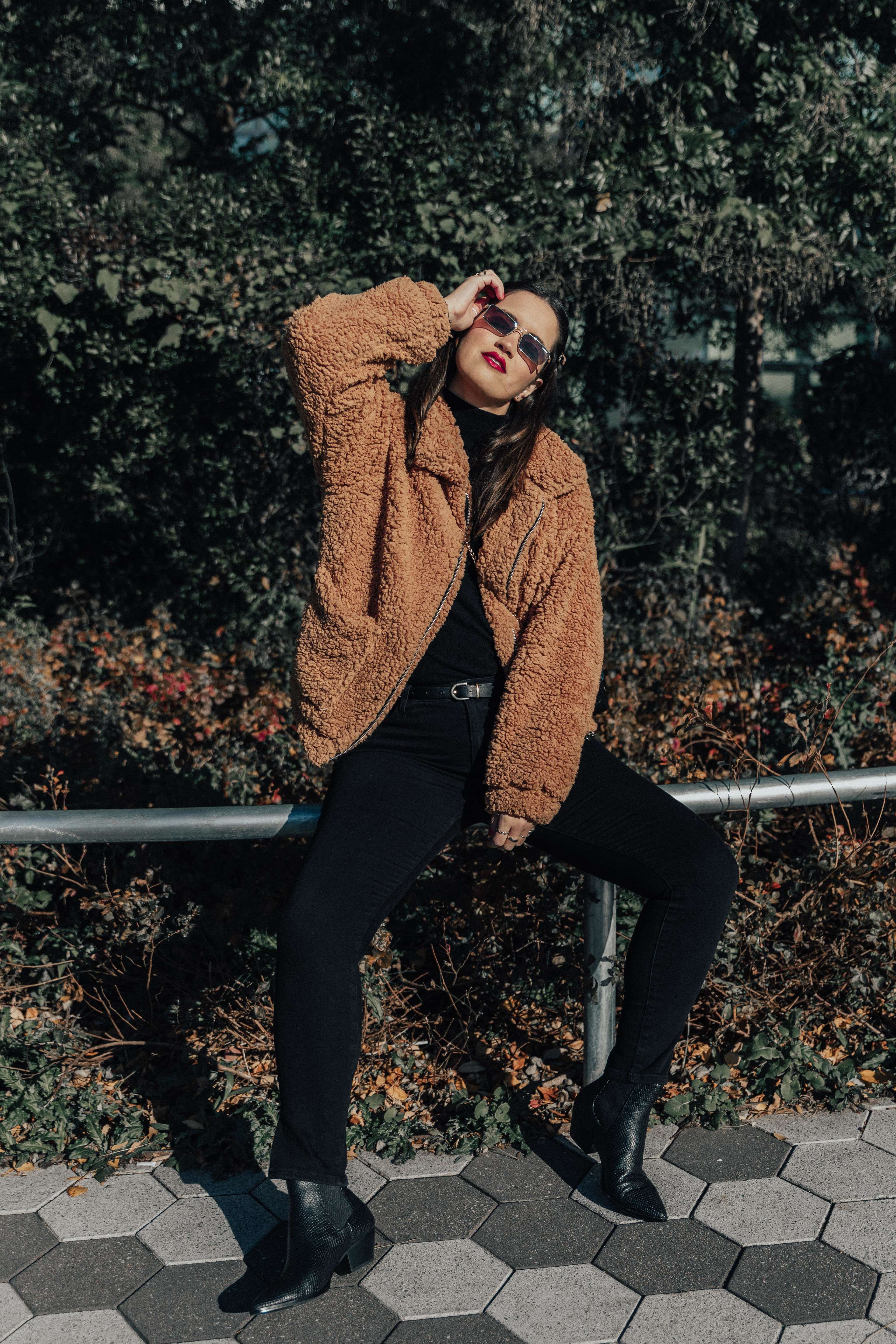 Woman in a cropped camel teddy jacket with black skinny jeans, ankle boots and sunglasses