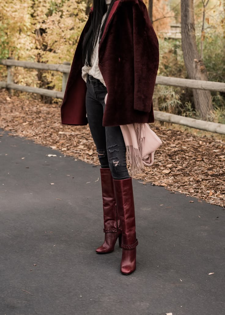 Woman wearing burgundy knee-high boots with black skinny jeans, a cream cable-knit sweater and a burgundy coat