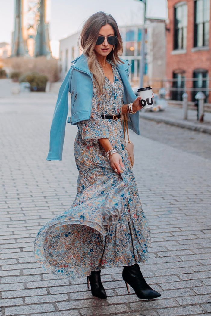 Woman in a floral maxi dress, blue moto jacket and black heeled ankle boots holding a coffee