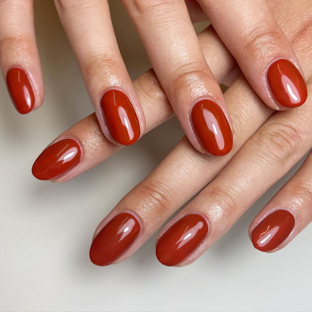 Short almond nails in a burnt terracotta rust-orange crème with a glossy finish; real photography.