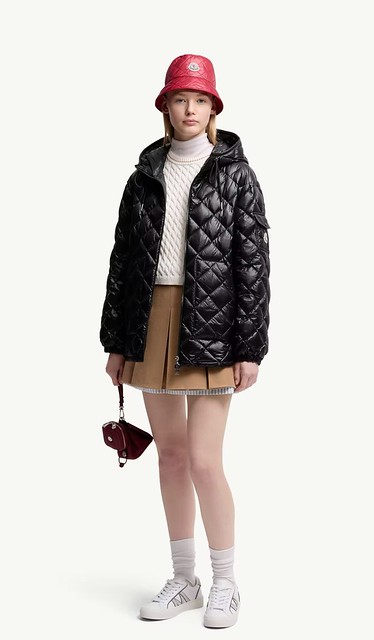 Woman in a black quilted jacket, cream turtleneck, tan pleated mini skirt, white socks and white sneakers with a red bucket hat