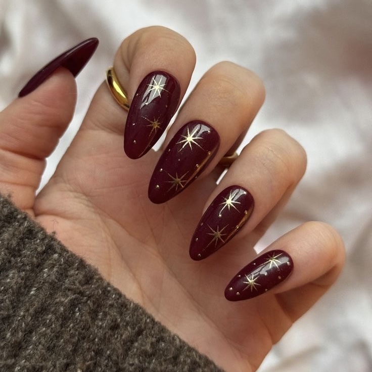 Glossy burgundy almond nails with scattered gold starburst nail art