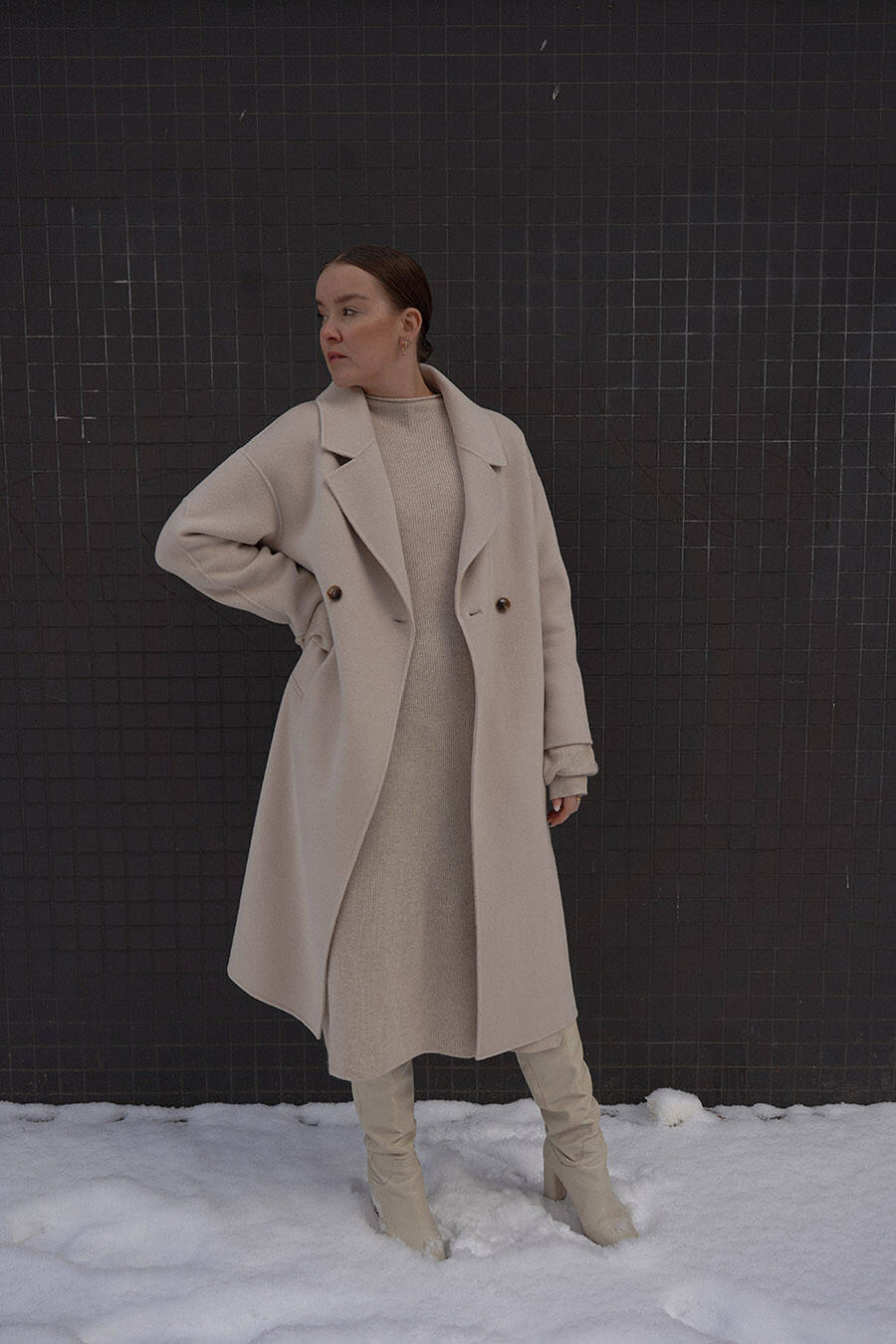 Woman in a cream monochrome winter outfit — long wool coat over a knit midi dress with knee-high boots in the snow
