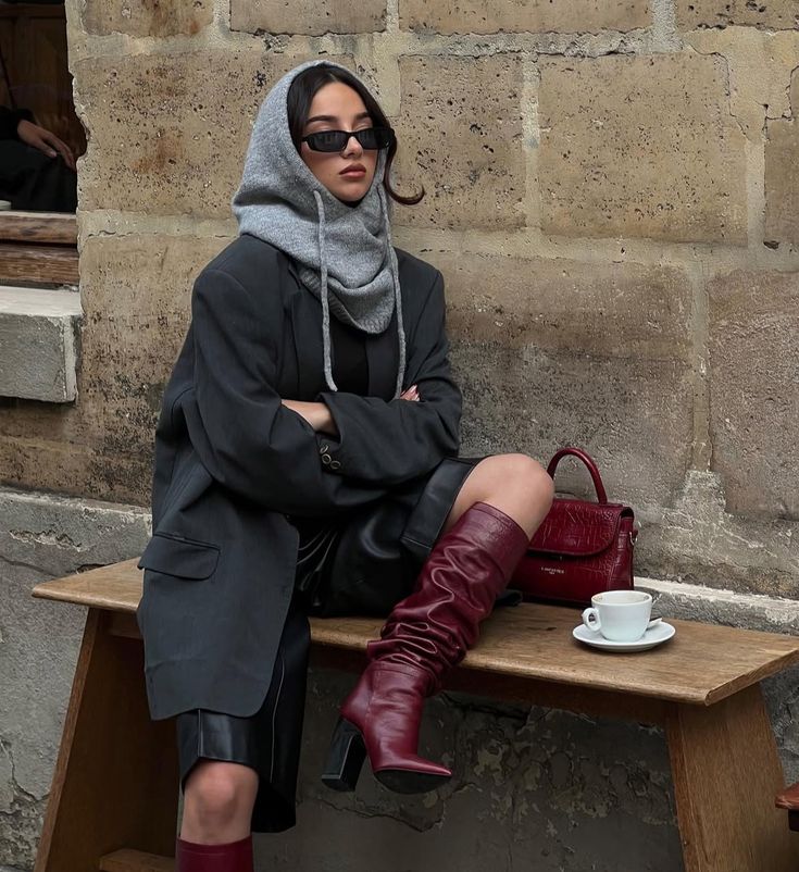 Woman seated in burgundy slouchy heeled boots with a black blazer, leather mini skirt and red mini bag