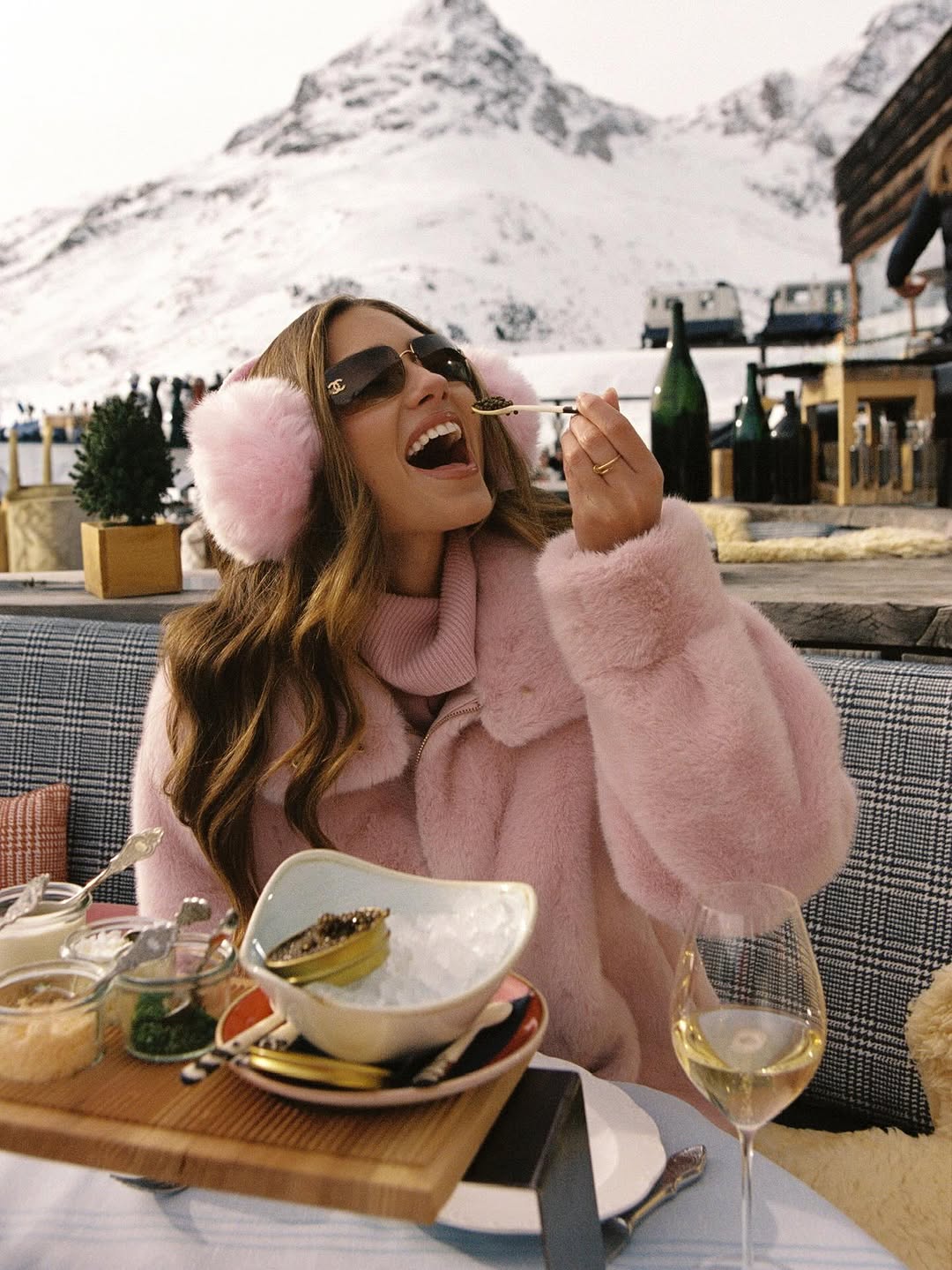 Woman in pink faux fur and pink earmuffs laughing over a meal at a snowy mountain restaurant