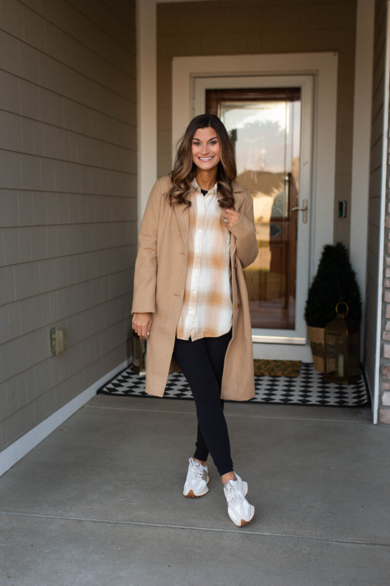 Woman in a tan coat with a beige plaid flannel shirt, black leggings and white sneakers