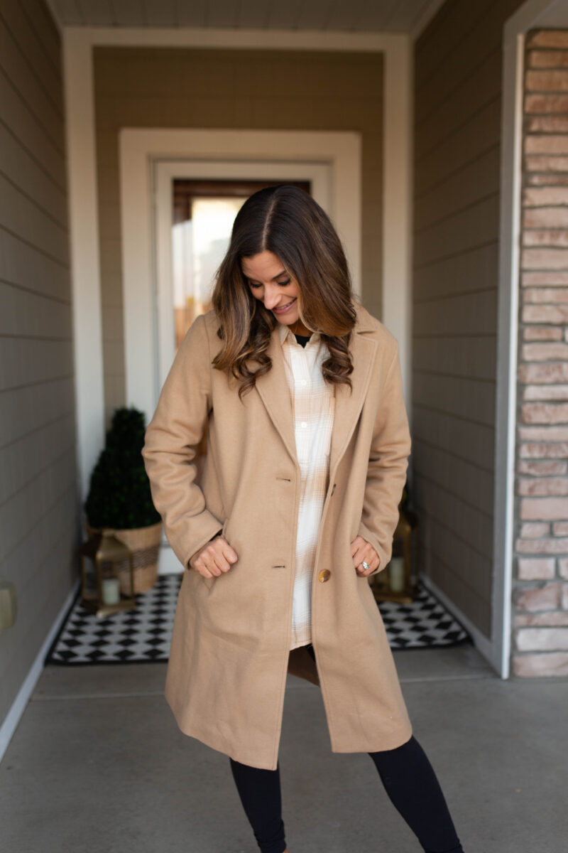 Woman in a tan coat over a white and tan plaid shirt with black leggings