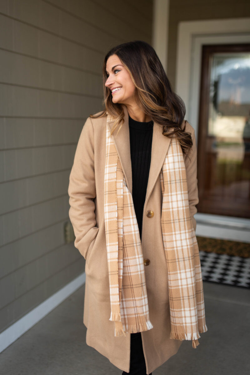 Woman in a tan coat with a camel plaid blanket scarf over a black top