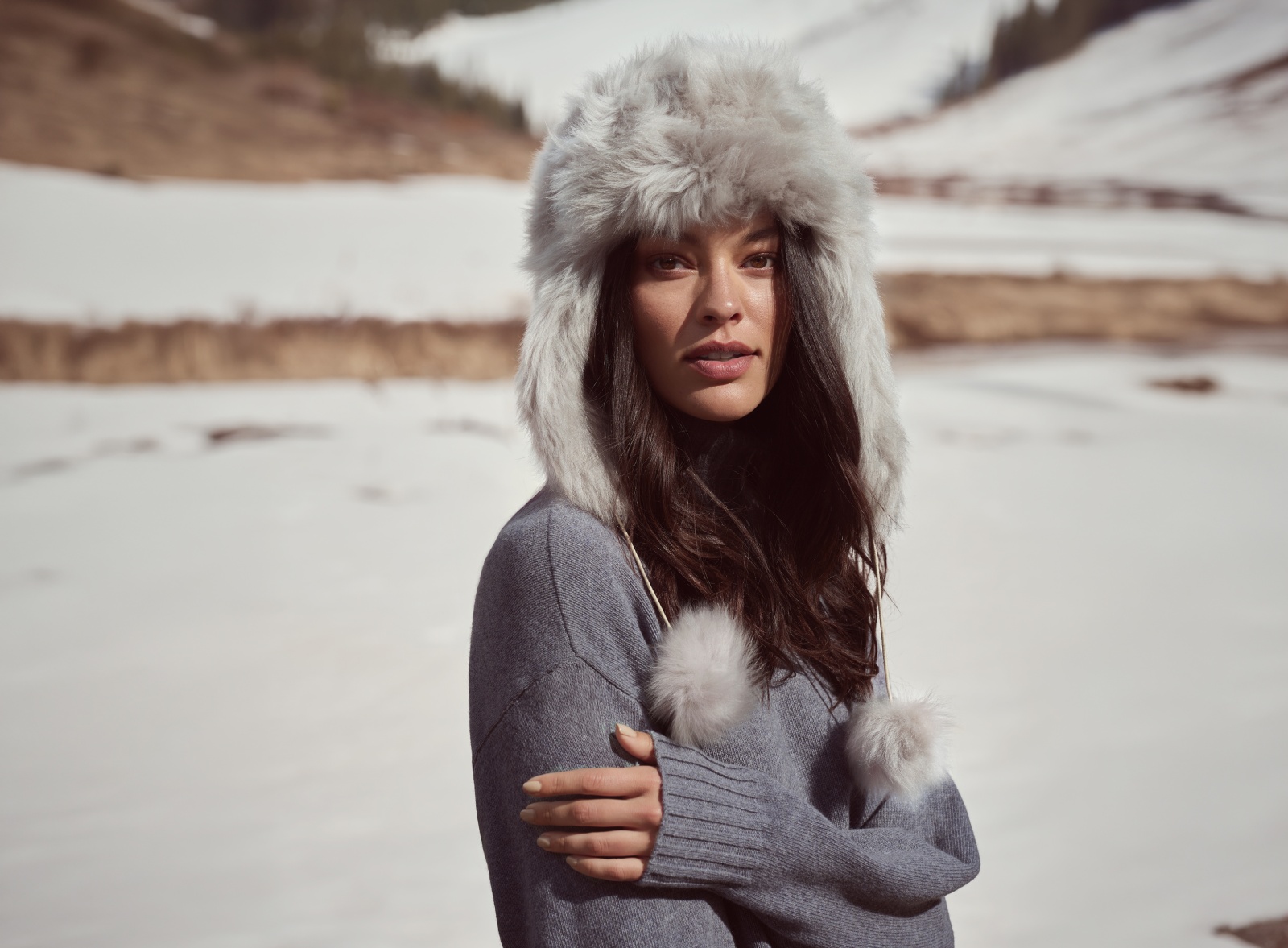 Woman in a grey faux fur trapper hat with pom-pom ties and a grey knit sweater in the snow