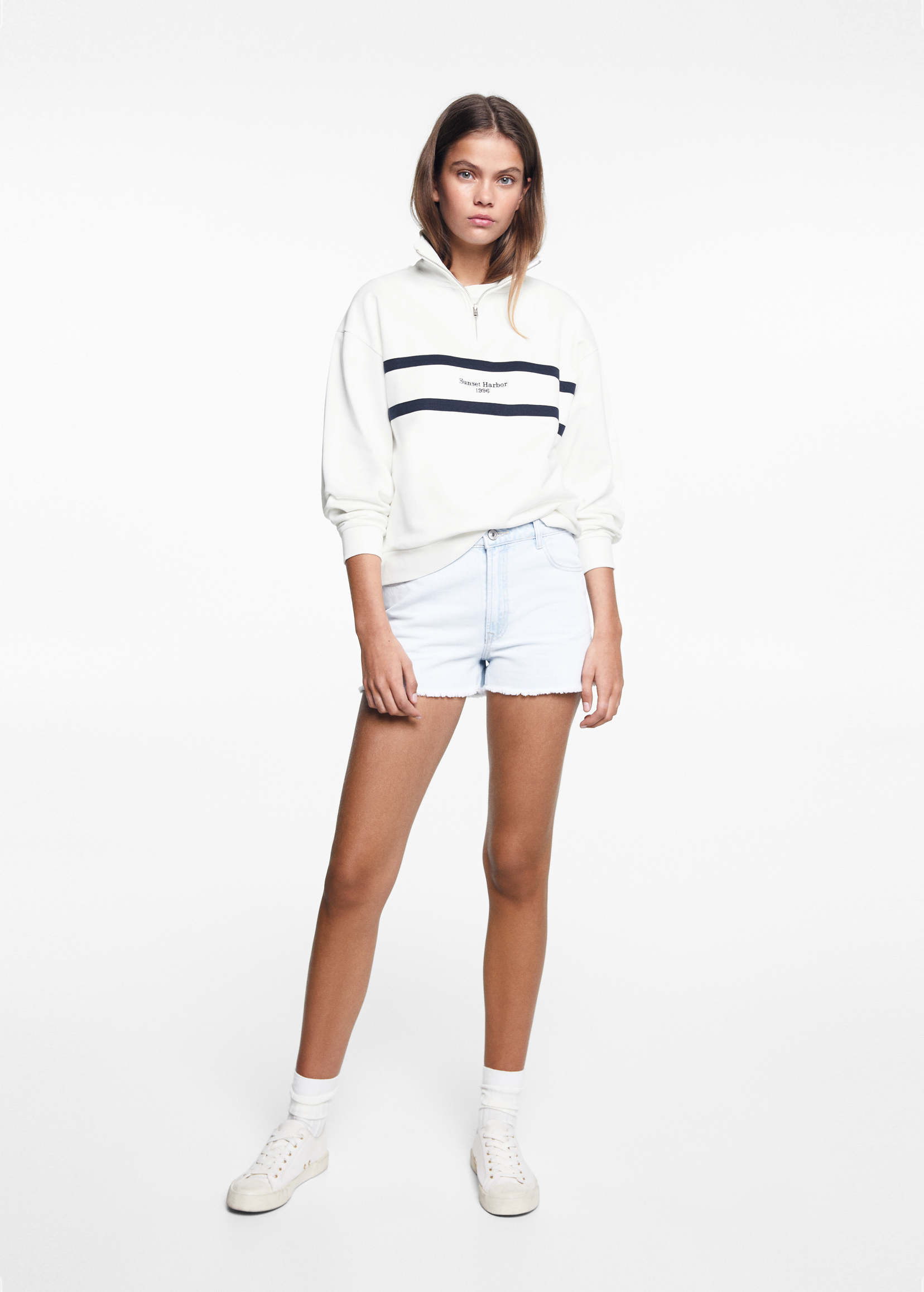 Woman in a white striped quarter-zip sweatshirt with white denim shorts and white sneakers