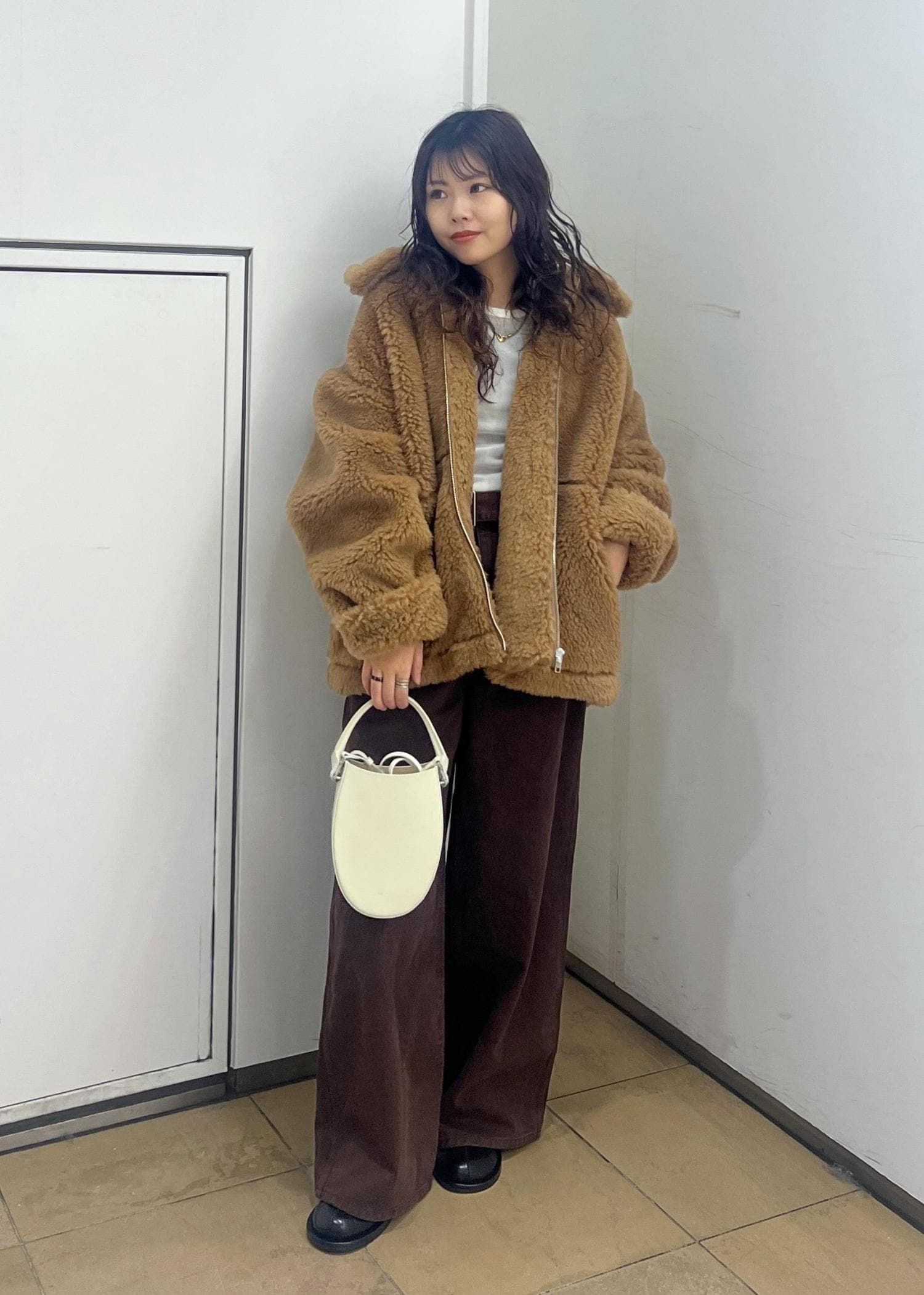 Woman in a caramel teddy fleece hooded jacket with dark wide-leg trousers and a white mini bag