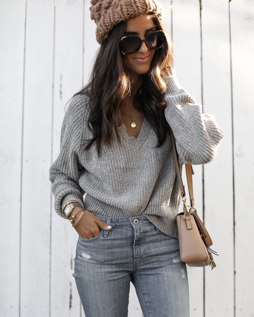 Woman in a tan pom beanie, grey sweater and grey jeans with sunglasses