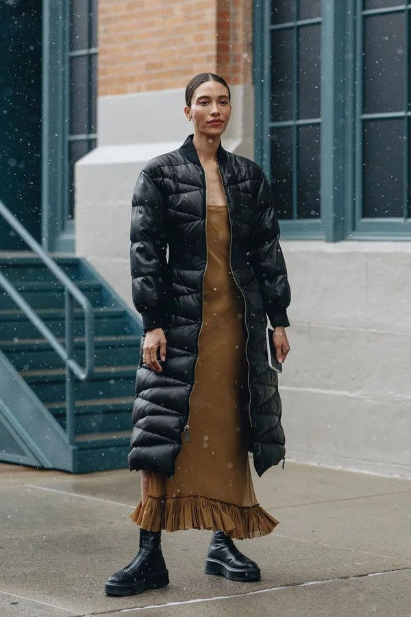 Woman in a long black puffer coat over a camel slip dress with chunky black boots in snow