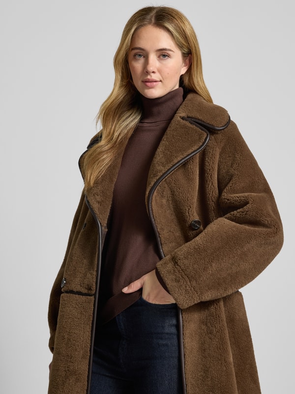 A mid-brown teddy coat with dark leather trim along the lapels, worn over a chocolate-brown turtleneck and dark indigo jeans.