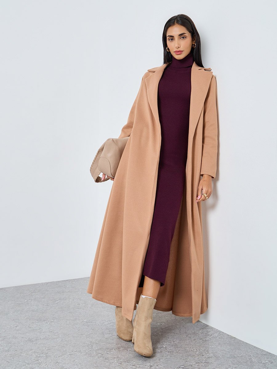 Woman wearing a long camel duster coat over a burgundy turtleneck midi dress with beige ankle boots