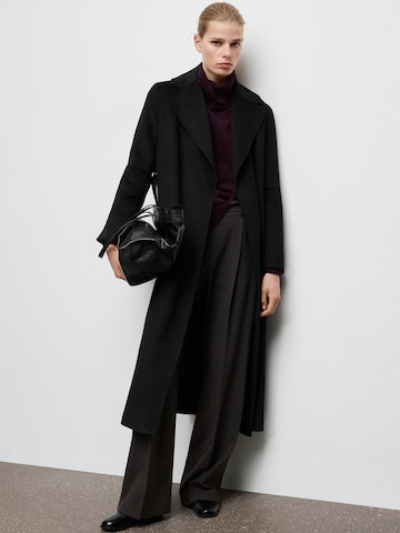 A black long coat worn with a burgundy turtleneck, dark wide trousers, loafers and a black bag.