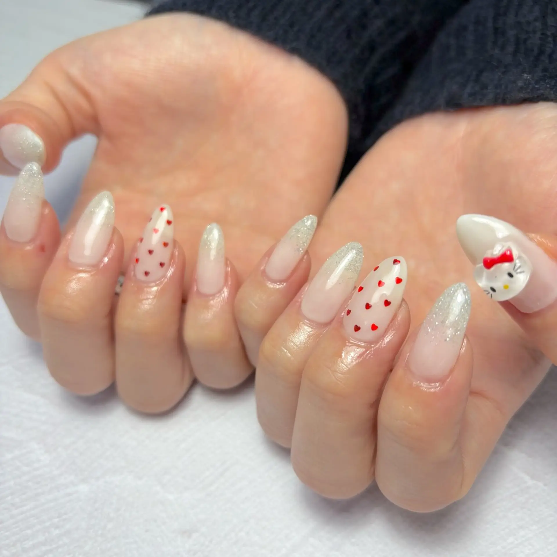Medium almond nails in milky white with silver glitter gradient tips, tiny red hearts and a Hello Kitty face with a red bow