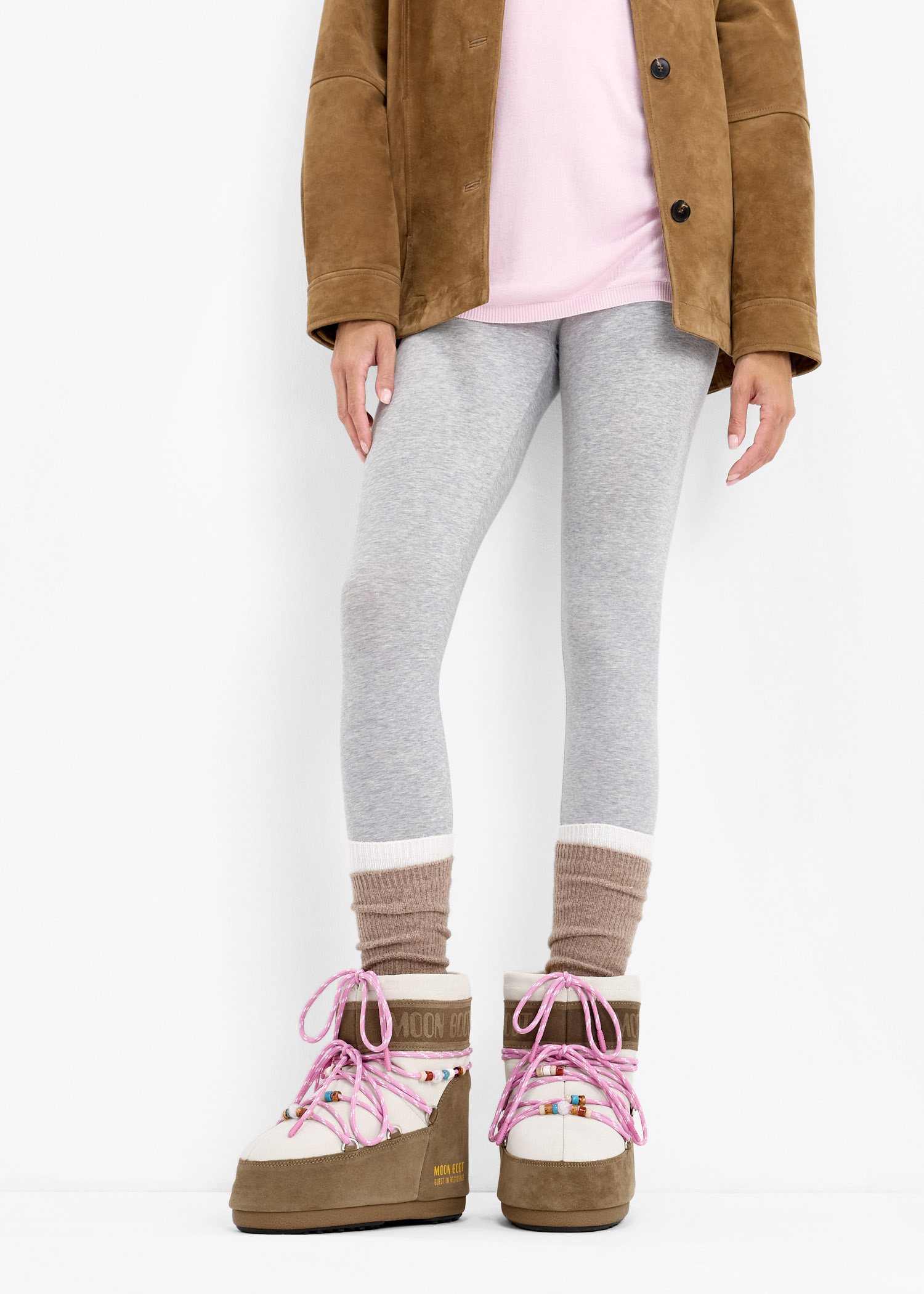 Woman in cream and brown suede Moon Boots with pink laces, grey leggings, pink top and brown suede jacket