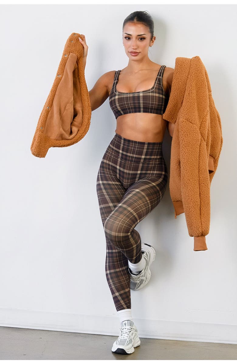 Woman in brown plaid sports bra and leggings set holding open a tan sherpa jacket, white sneakers